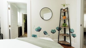 A bedroom with a bed, a wall-mounted mirror, a shelf with plants, and floral wall art.