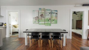 A dining area with a wooden table and black chairs, a white wall with a painting, and a kitchen in the background.