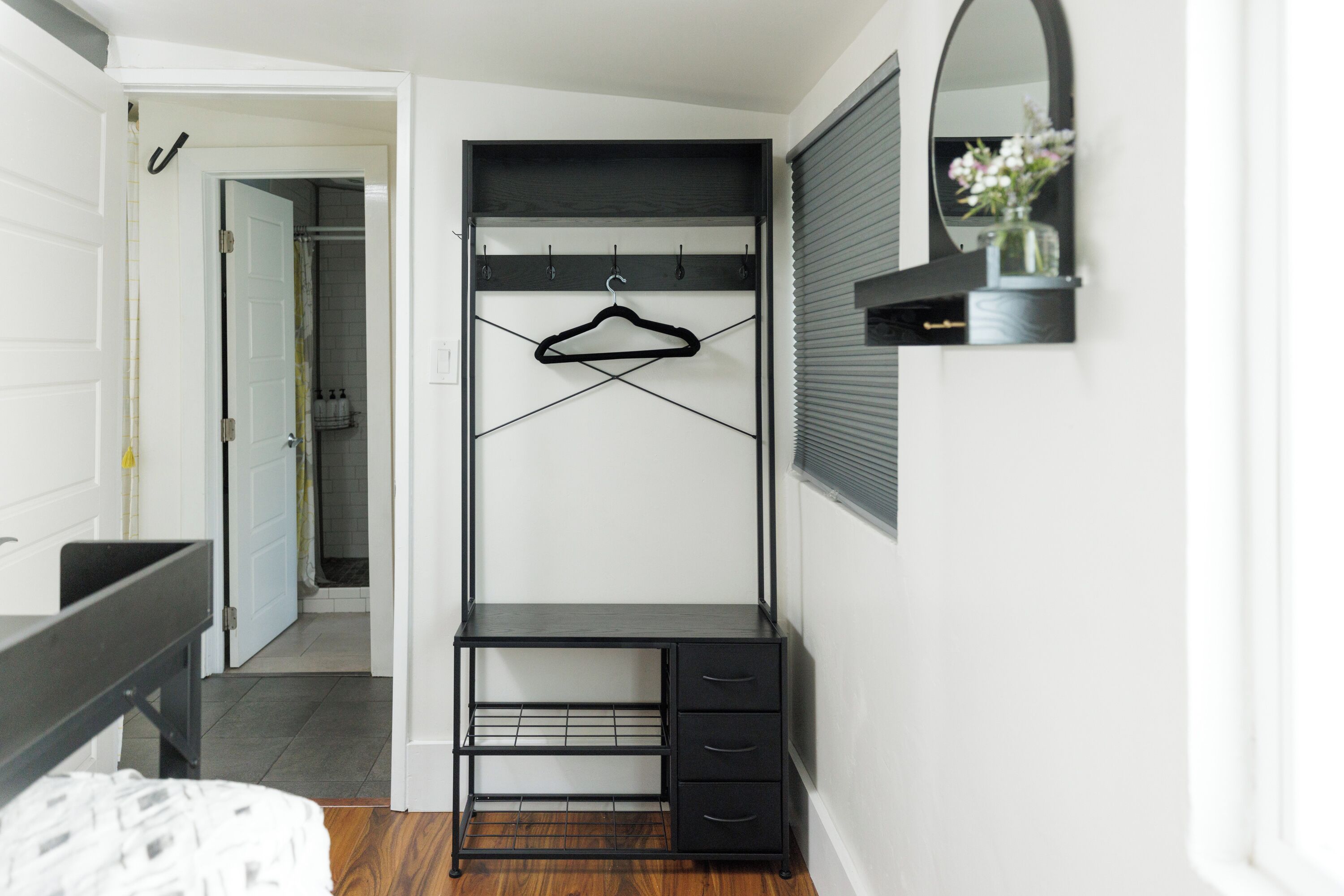 A compact bedroom with a bed, a small desk with drawers, a hanging rack, and a mirror.