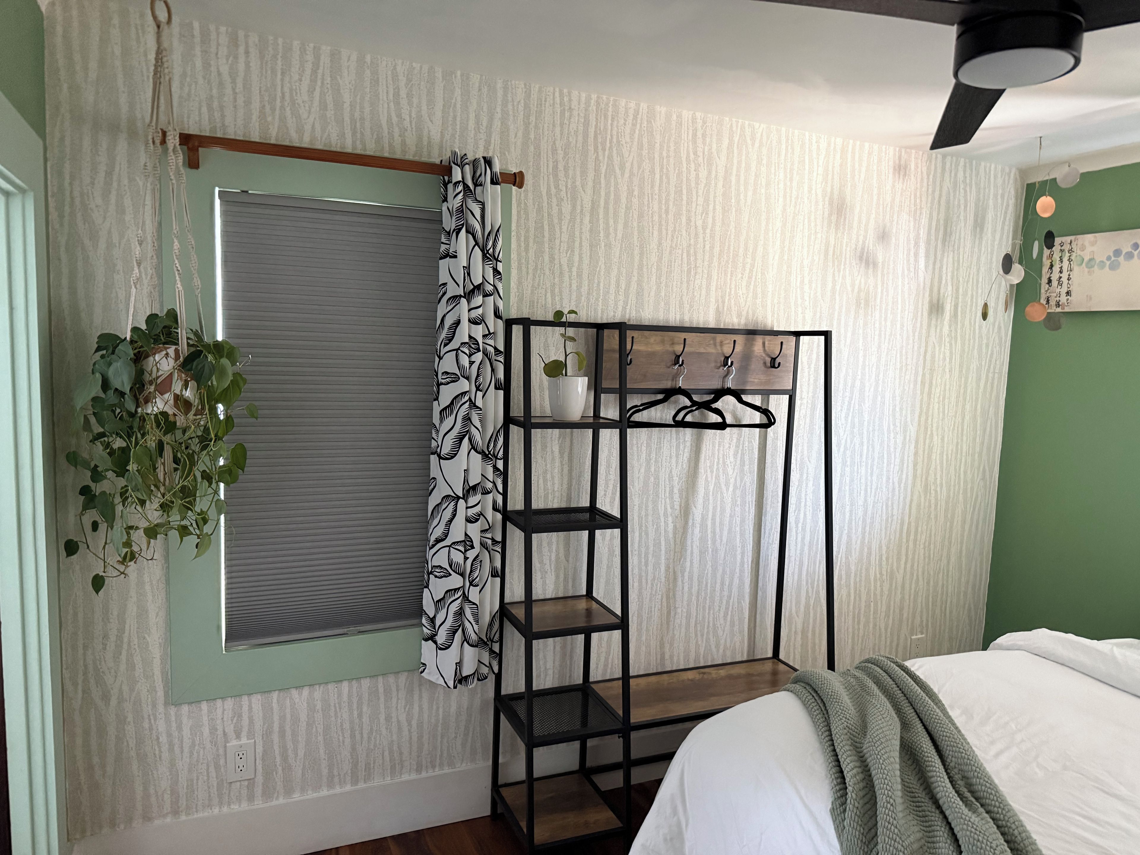 Een slaapkamer met een bed, een raam met gordijn, een wandplank met haken en een hangplant.