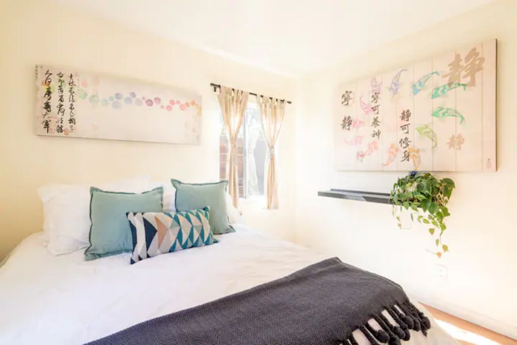 Een slaapkamer met een bed, kussens, een deken, een raam met gordijnen en wanddecoratie.