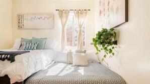 Un dormitorio con cama, almohadas, una planta en la mesita de noche y una ventana con cortinas.