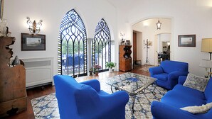 A living room with blue armchairs, a glass coffee table, and a patterned rug. There are two stained glass windows and a wooden cabinet with decorative items. - AMORE RENTALS - Villa Angelo with Private Terraces, Parking and Air Conditioning (Sant´Agnello)