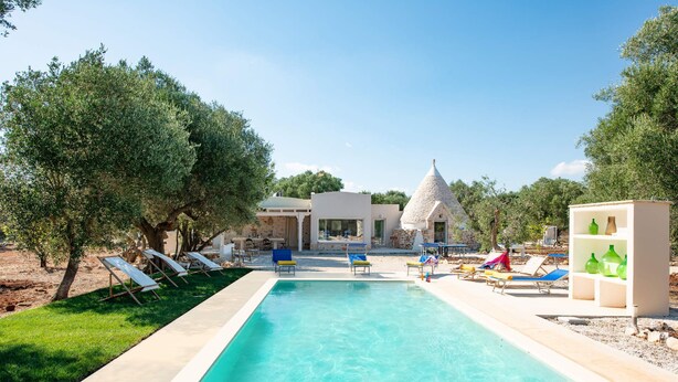 Un espace piscine avec des chaises longues, un bâtiment en pierre surmonté d’un toit conique et un ciel d’un bleu limpide.