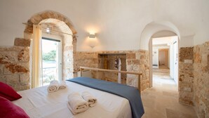 Une chambre avec des murs en pierre, un lit recouvert de draps blancs et une vue sur l’extérieur.