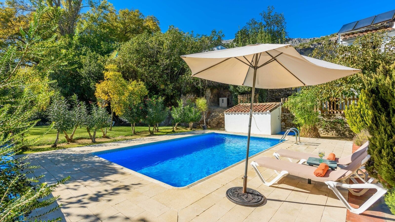 Une piscine rectangulaire à l’eau d’un bleu limpide, entourée d’une terrasse dallée. On y trouve des chaises longues et un parasol pour se protéger du soleil.