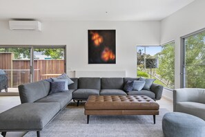 A modern living room with a sectional sofa, a leather ottoman, and a large window offering a view of the outdoors. - Seahaven - Brilliant Location, Stunning Modern Features (Lorne)