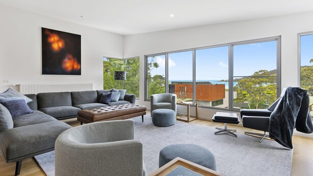 A modern living room with a grey sofa, ottoman, and armchairs, a large window with a view of the sea, and a painting on the wall. - Seahaven - Brilliant Location, Stunning Modern Features (Lorne)