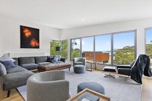 A modern living room with a grey sofa, ottoman, and armchairs, a large window with a view of the sea, and a painting on the wall. - Seahaven - Brilliant Location, Stunning Modern Features (Lorne)
