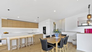 A modern kitchen with a central island, wooden flooring, and a dining area with a table and chairs. - Seahaven - Brilliant Location, Stunning Modern Features (Lorne)
