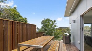 A wooden deck with a metal-framed table and chairs, overlooking a residential area with trees and a hill in the background. - Seahaven - Brilliant Location, Stunning Modern Features (Lorne)