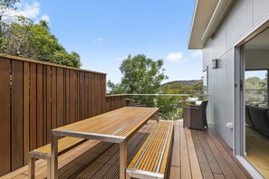 A wooden deck with a metal-framed table and chairs, overlooking a residential area with trees and a hill in the background. - Seahaven - Brilliant Location, Stunning Modern Features (Lorne)