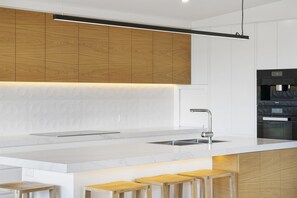 Una cocina moderna con encimera blanca, armarios de madera, un lavamanos de acero inoxidable y un horno empotrado.