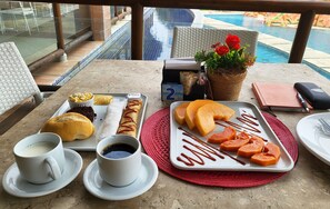Una mesa con diversos alimentos, como pasteles, una bandeja de fruta cortada y una rebanada de pan con mermelada. También hay dos tazas de café y un cuaderno con un bolígrafo sobre la mesa.