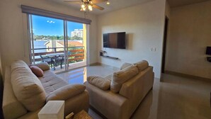 A spacious living room with a large sliding glass door leading to a balcony, a flat-screen TV mounted on the wall, and a ceiling fan.