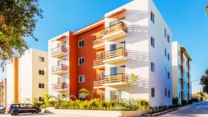 A multi-story residential building with balconies, a car parked in front, and a clear sky.