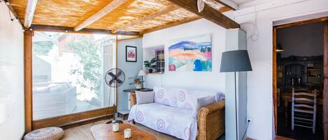 A cozy room with a wicker sofa, a wooden coffee table, a standing fan, and a large window offering a view of the outdoors.