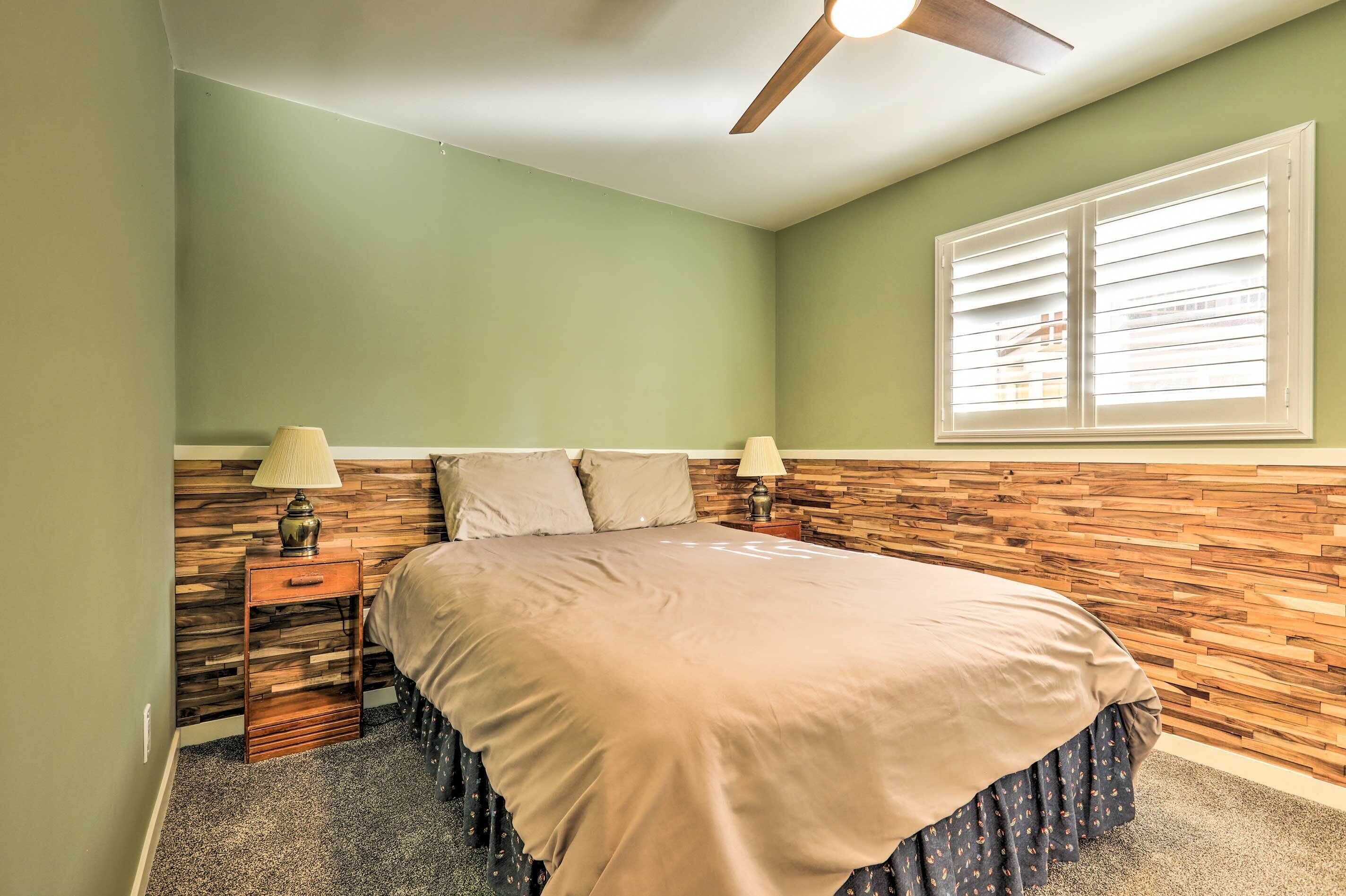 A bedroom with a bed, two bedside lamps, a wooden headboard, a ceiling fan, and a window with blinds.