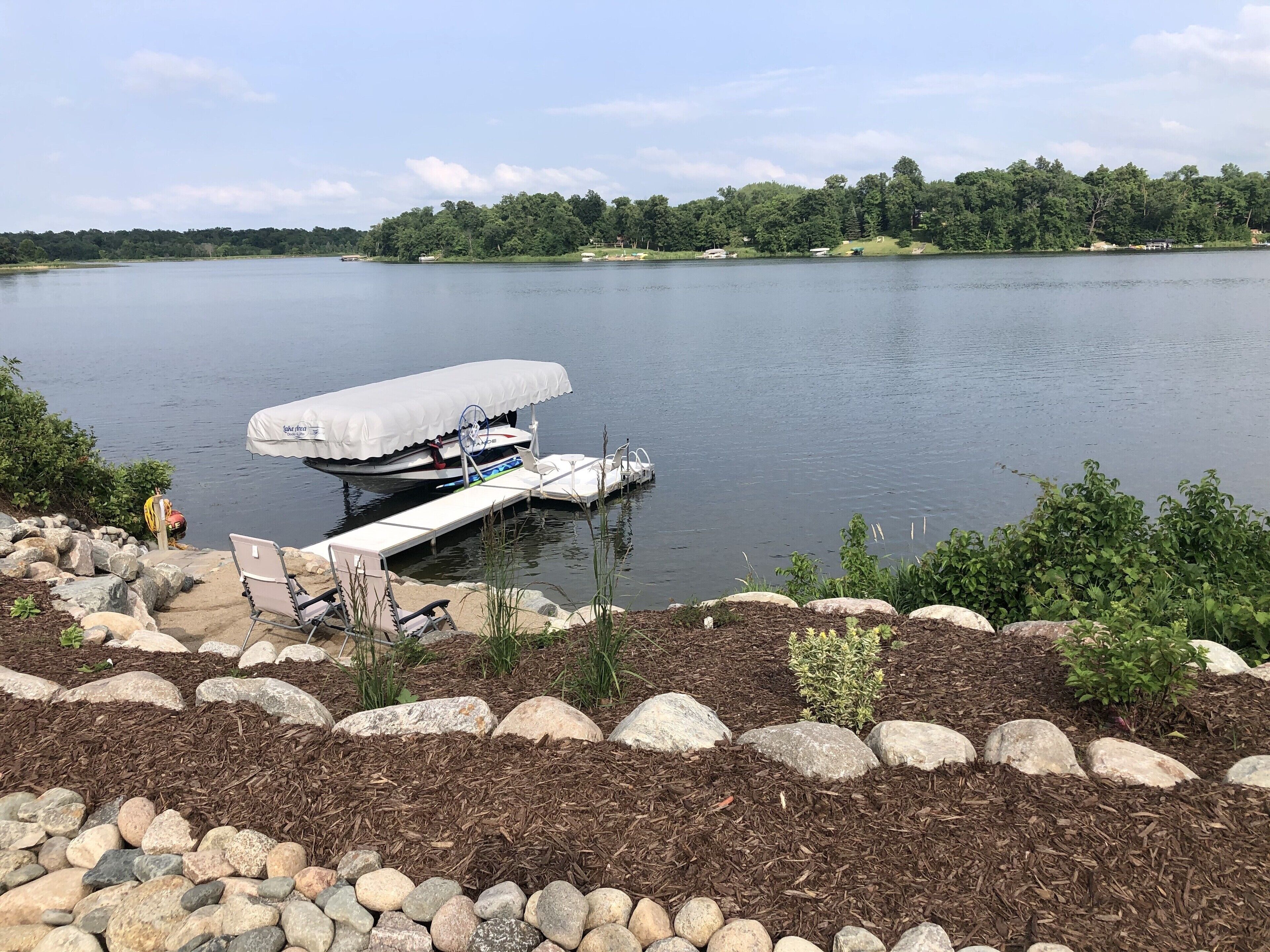 A dock with a covered boat, a lounge chair, and a rocky shoreline with mulch and plants.