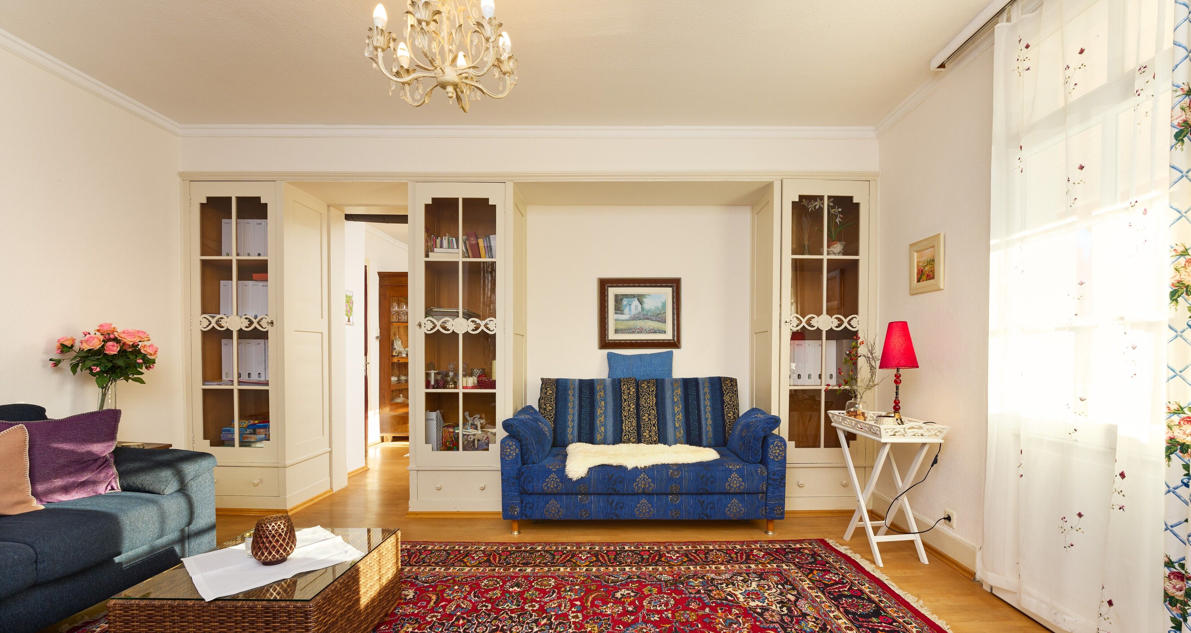 A living room with a blue sofa, a red patterned rug, a chandelier, and a white cabinet with glass doors.