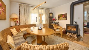 Een gezellige woonkamer met een ronde houten tafel, rieten meubels, een open haard en een groot raam met gordijnen.