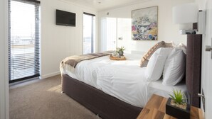Een moderne slaapkamer met een groot bed, een flatscreen televisie, een schilderij en een raam met luxaflex.