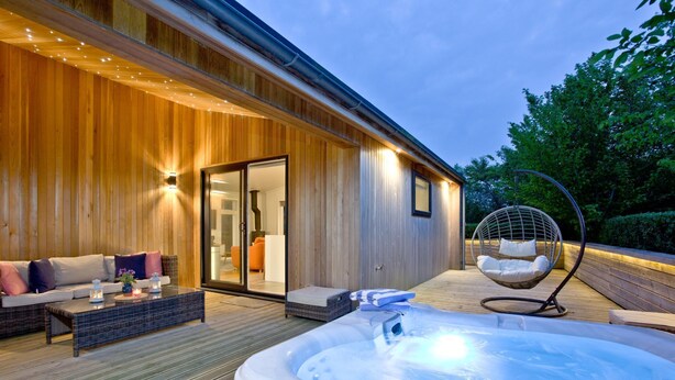 A wooden exterior of a building with a hot tub, a hanging chair, and a sofa with cushions.