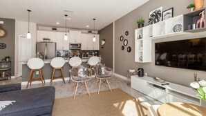 A modern kitchen with white cabinets, a central island, and pendant lighting. There is a dining area with a white table and chairs, and a living room with a sofa and a TV. - Storey Lake - LUXURY 4 bedrooms, 3 bath, private pool -  Star Wars & Mickey theme (Kissimmee)