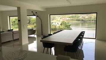 A modern dining area with a large table and chairs, a sofa, and a view of a lake and buildings.