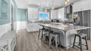 A modern kitchen with white cabinets, a large island with bar stools, stainless steel appliances, and pendant lighting. - Luna's: Amazing Ocean View House with Pool, Spa, Elevator and Electric Car Charg (Oak Island)