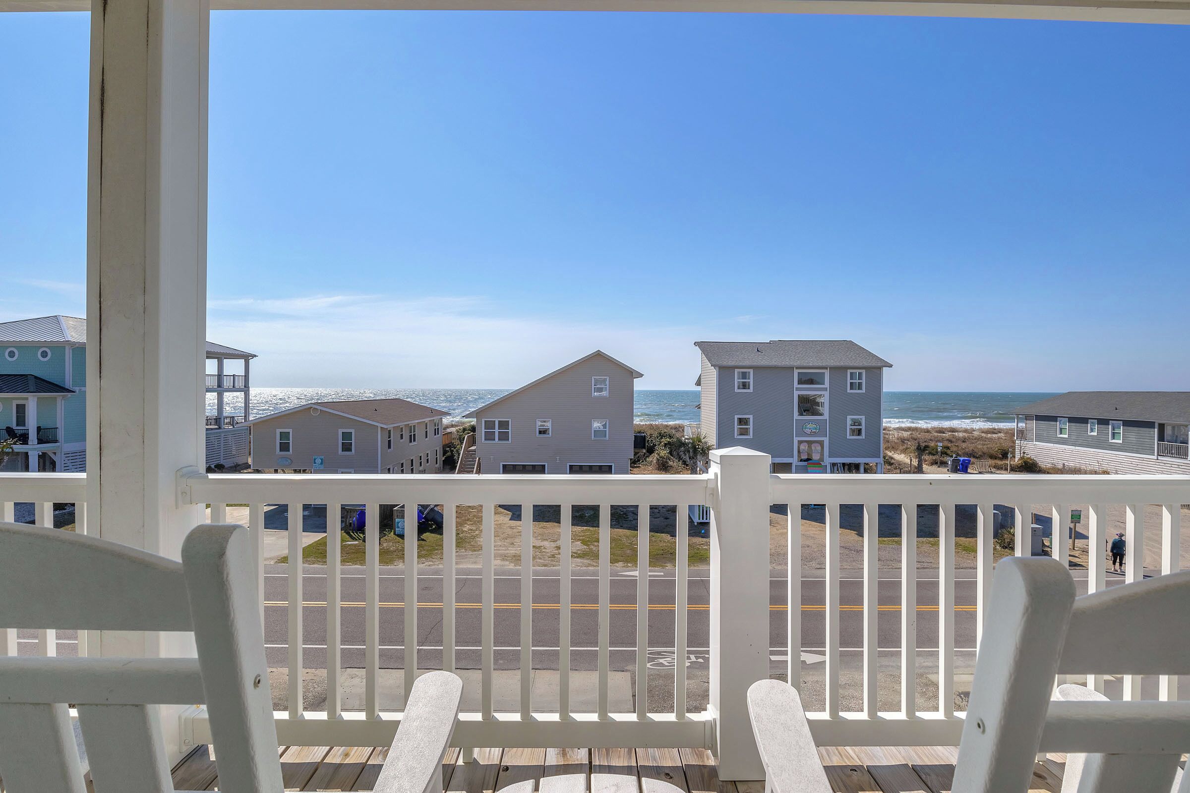 Ein Balkon mit weißen Geländern, der Blick auf einen Strand mit Häusern im Hintergrund freigibt.