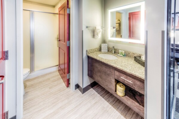 A modern bathroom with a shower, a sink with a granite countertop, and a mirror with a light above it.