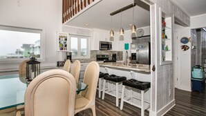 A modern kitchen with white cabinets, stainless steel appliances, and a glass dining table with four chairs. - Out Of Office: Dog-friendly, Oceanfront Home on West Beach with Corn Hole and Ho (Oak Island)