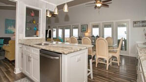 A kitchen with a central island, white cabinets, and a dishwasher. There is a dining area with a table and chairs. - Out Of Office: Dog-friendly, Oceanfront Home on West Beach with Corn Hole and Ho (Oak Island)