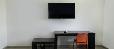 A modern, minimalist room with a flat-screen TV mounted on the wall, a black desk with a chair, and a small bench.