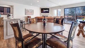 Un comedor moderno con una mesa redonda y sillas, una pared de piedra con un logo, y un ventanal con vistas a árboles.