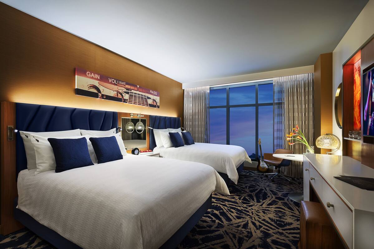 A hotel room with two beds, a desk, a TV, and a large window with curtains.
