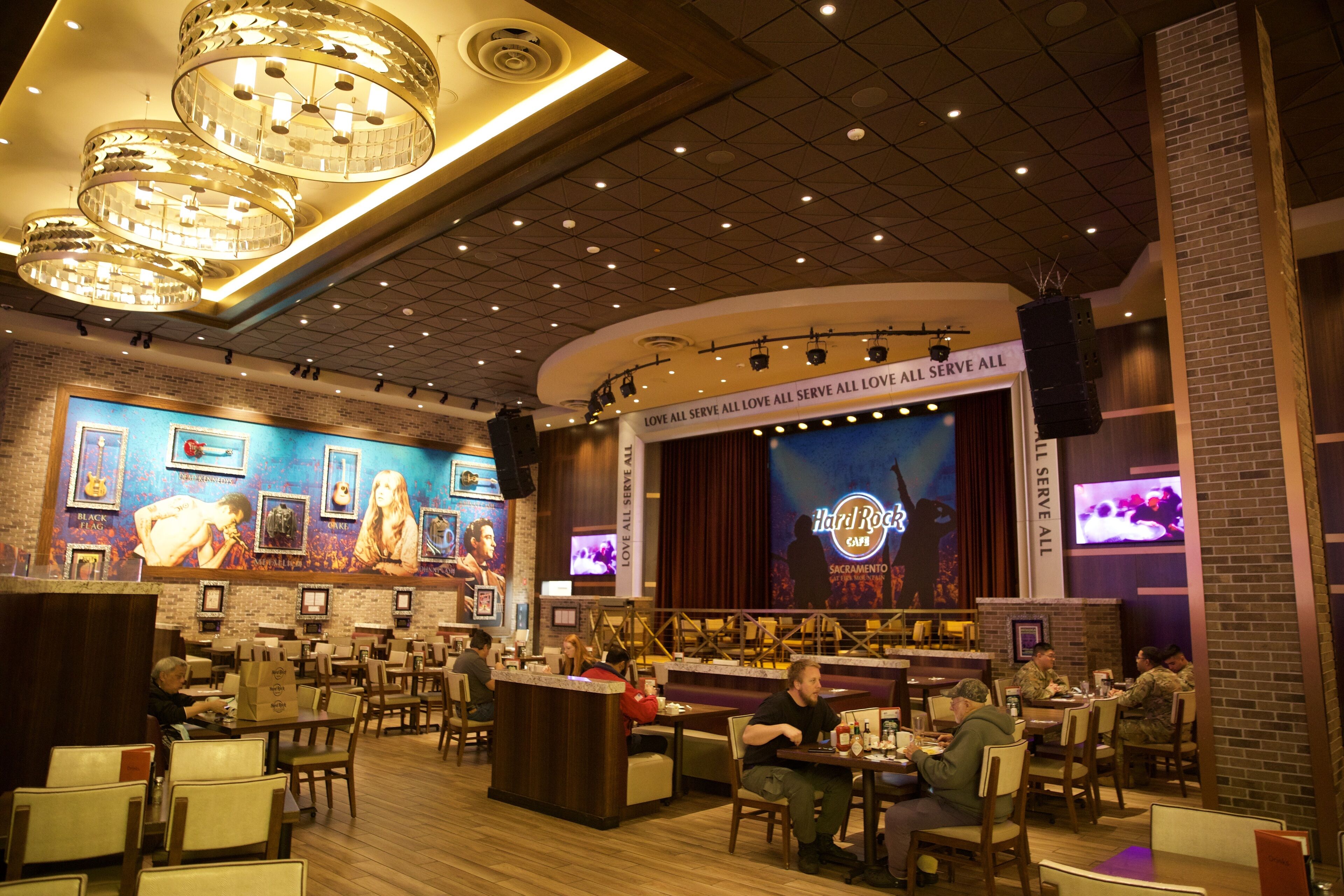 A restaurant with a stage and a large screen displaying 'Hardi Rock Cafe' and 'Sacramento'. There are tables and chairs arranged for dining.