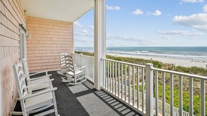 A balcony with white rocking chairs, a view of the beach, and a clear sky.