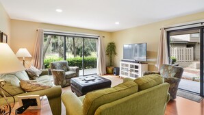 Een woonkamer met een bank, fauteuil en poef, een televisie op een kast, en een grote schuifpui naar een tuin.