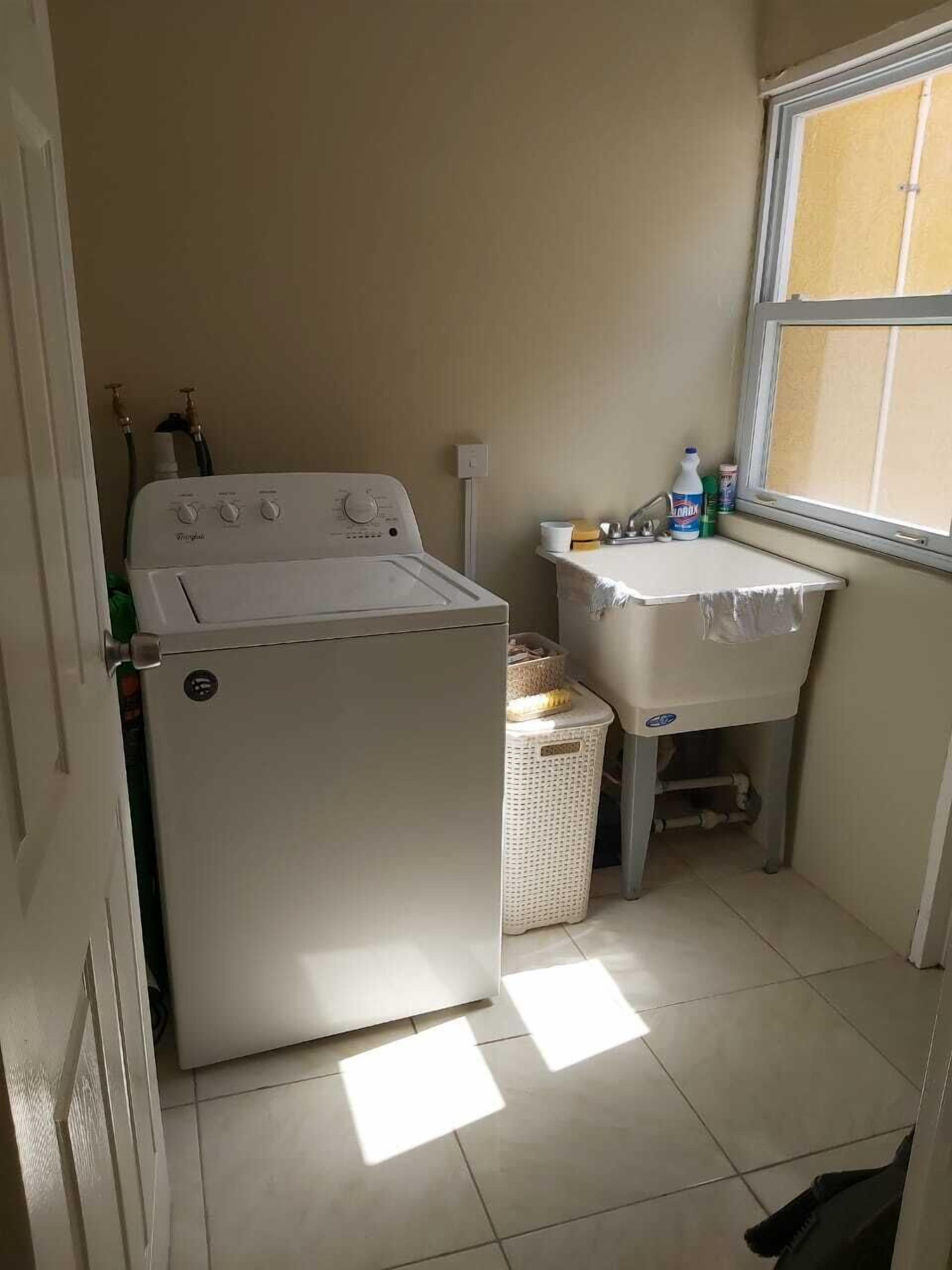 A compact laundry room with a front-loading washer and dryer, a small sink with a window above, and a basket on the floor.