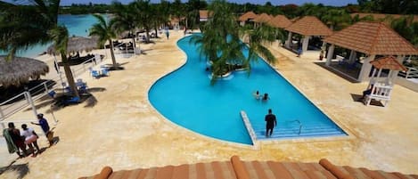 Un área de piscina en un resort con una piscina curvada, palmeras y estructuras de techo de paja.