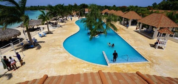 Un área de piscina en un resort con una piscina curvada, palmeras y estructuras de techo de paja.