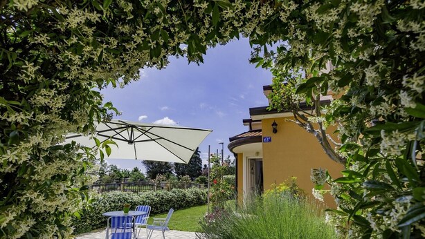 A patio area with stone flooring, blue striped chairs, a white umbrella, and a yellow building with a sign displaying '63'. - Apartment in a quiet location (Umag)