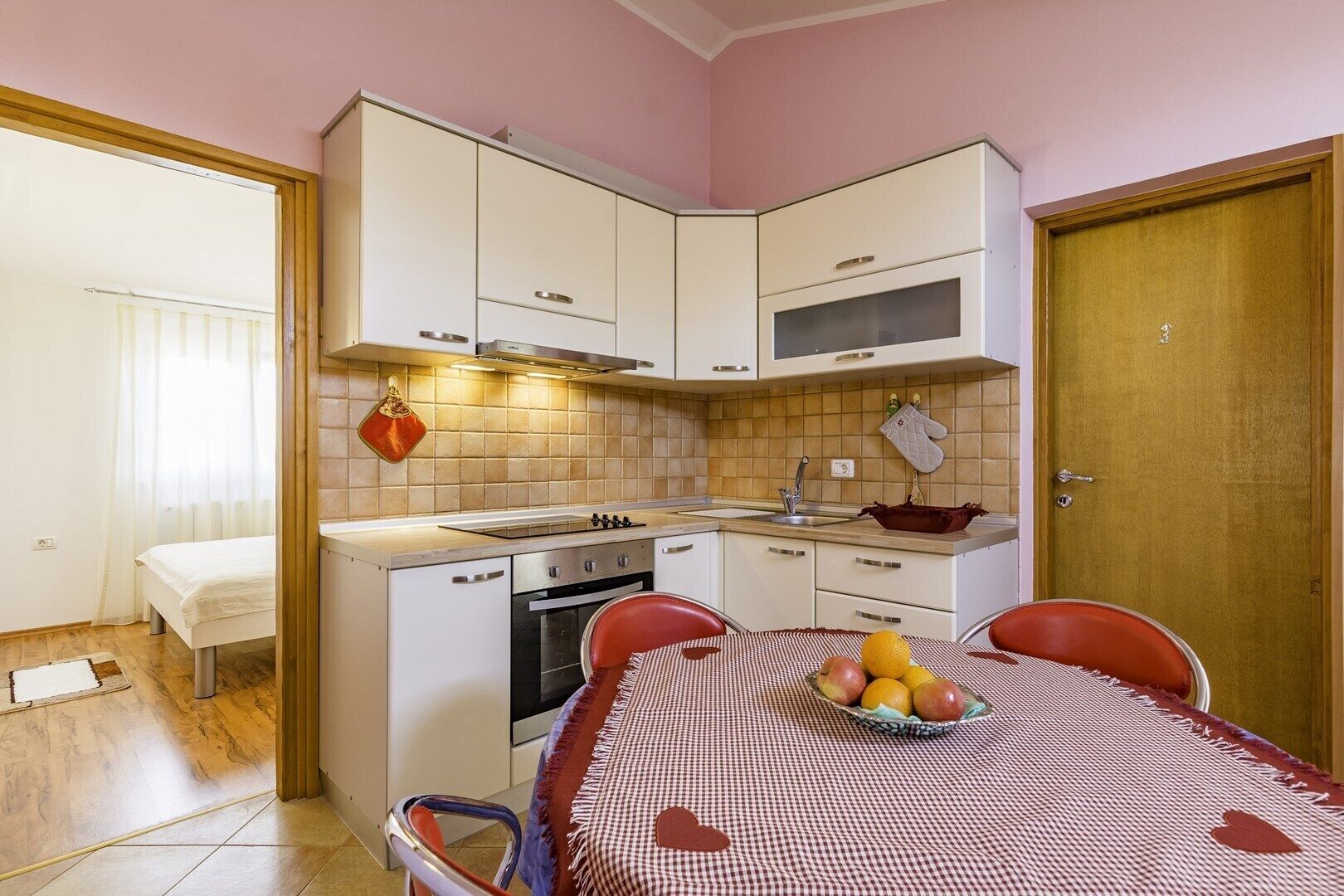 A compact kitchen with white cabinets, a stove, oven, and a sink. A dining table with a checkered tablecloth and a bowl of fruit is in the center.