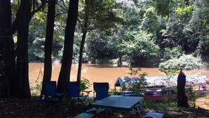 Un espacio de descanso a la orilla del río con mesas y sillas, rodeado de árboles y vistas a un río embarrado.