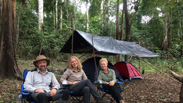 Tres personas sentadas en sillas de camping bajo una tienda de campaña en un bosque.