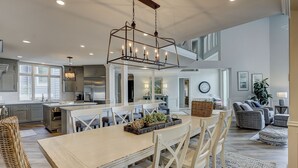 Una cocina amplia con una isla central, electrodomésticos modernos y una zona de comedor con sillas blancas y una mesa con camino de mesa tejido.