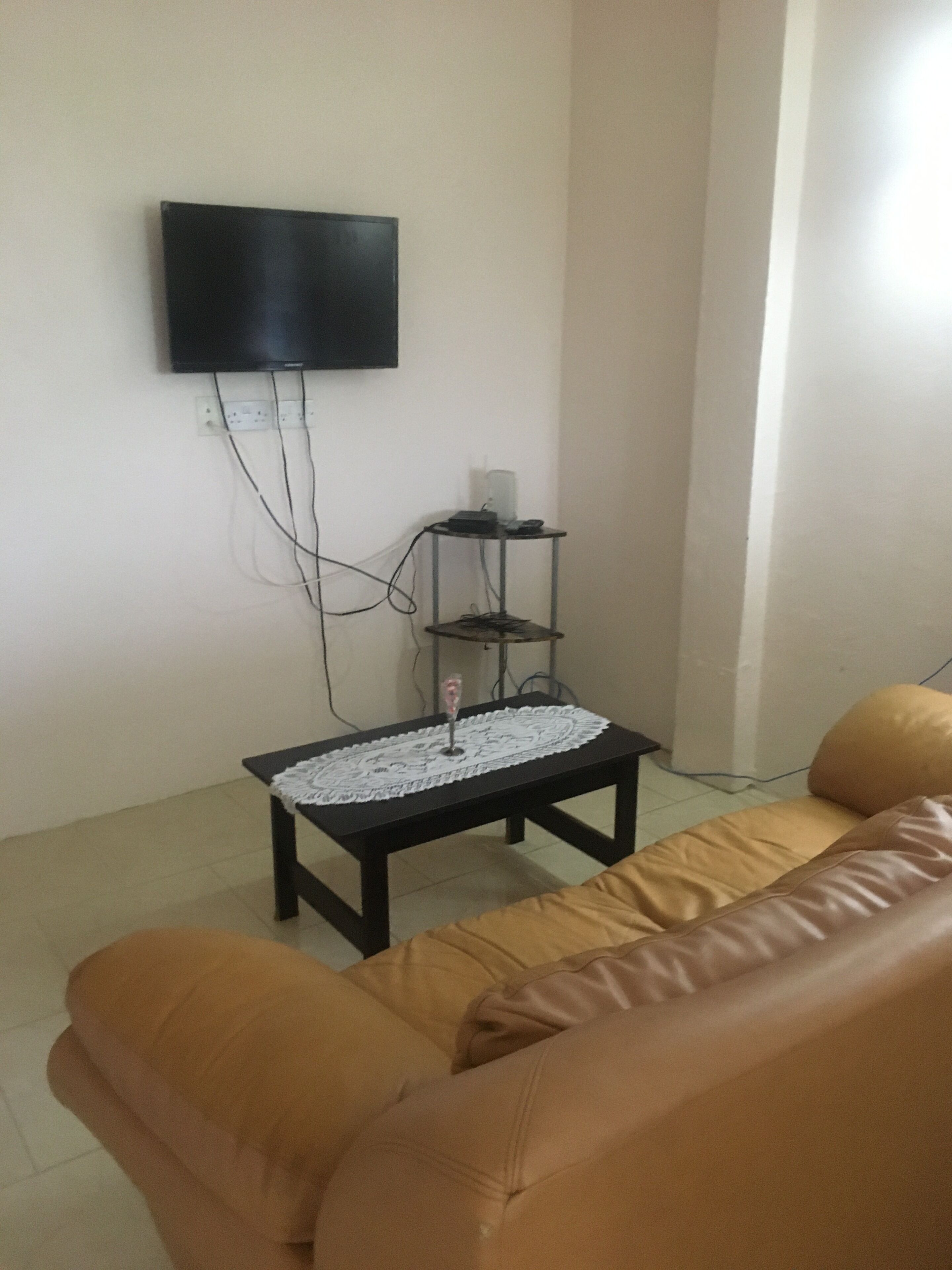 A living room with a brown leather sofa, a small black coffee table with a lace table runner, a flat-screen TV mounted on the wall, and a white wall socket.