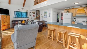A spacious living area with a high ceiling, wooden beams, a large chandelier, a dining table with stools, a kitchen with modern appliances, and a cozy seating area with a fireplace. - Cabin on the River - In Town on the Chattahoochee River (Helen)